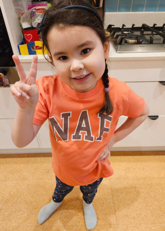 Child in an orange NAP T-shirt making a peace sign in a kitchen.