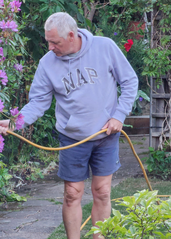 Person wearing a lavendar NAP hoodie, using a garden hose in a garden setting with plants and flowers.